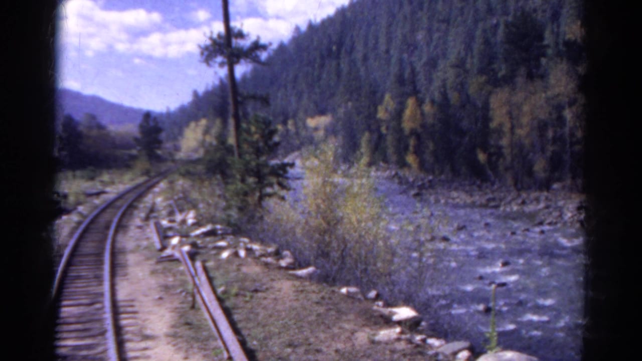 Curving railroad tracks run beside a river in a wooded mountain valley.