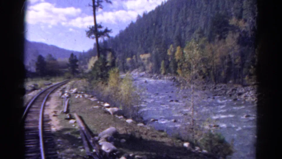 1967 Colorado 8mm frame from a moving train, showing rails, river water, rocks, trees, and a mountain slope in daylight.