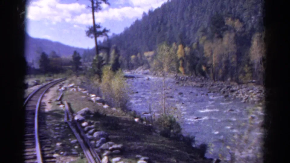 1967 Colorado home-movie frame showing tracks bending beside a rushing river, with forested mountains and autumn trees beyond.