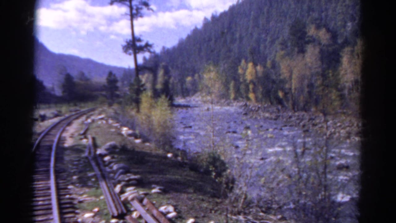 1967 Colorado 8mm frame showing railroad tracks curving beside a river, with forested mountain slopes and blue sky beyond.
