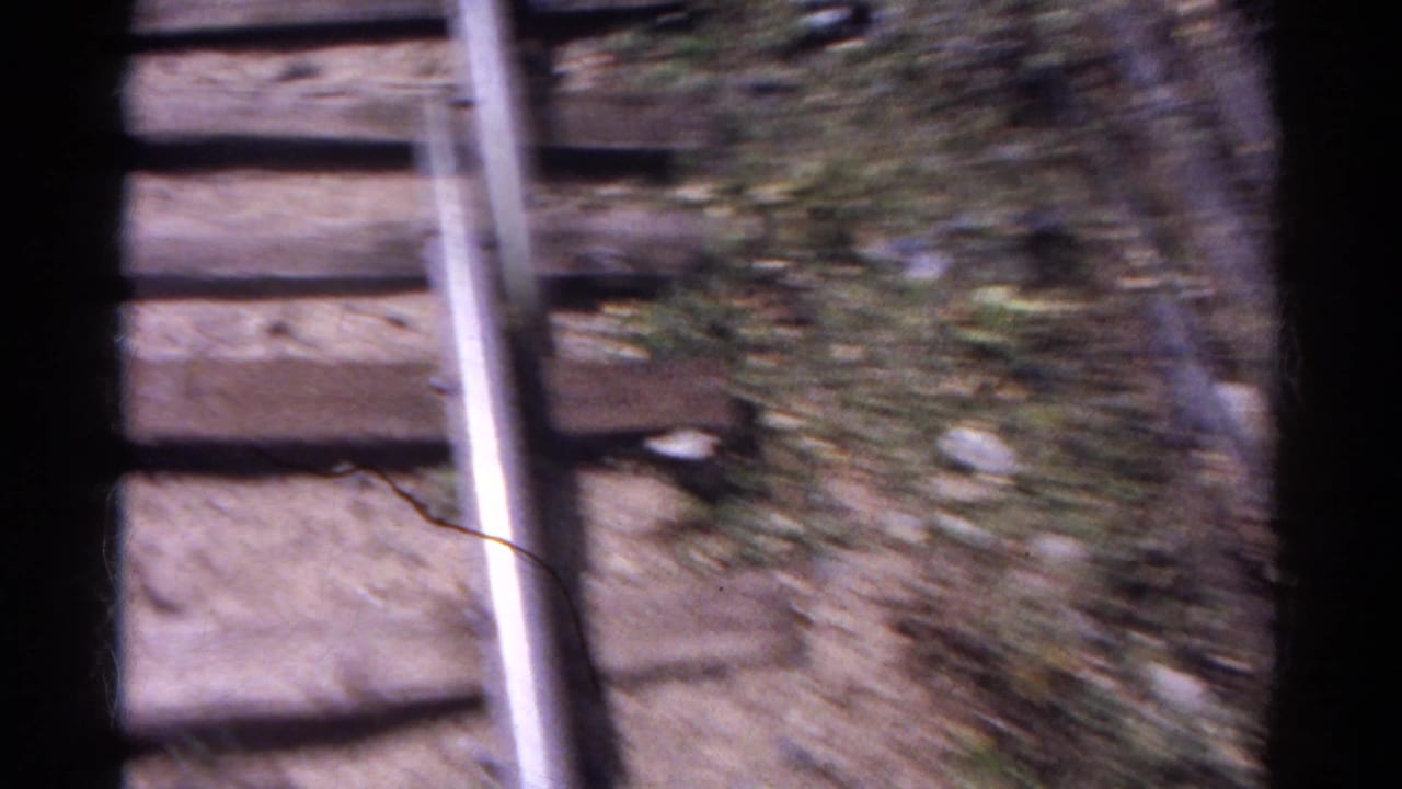 Close rear-facing train view of wooden ties, a rail, and blurred trackside ground during motion in the Colorado mountain sequence.