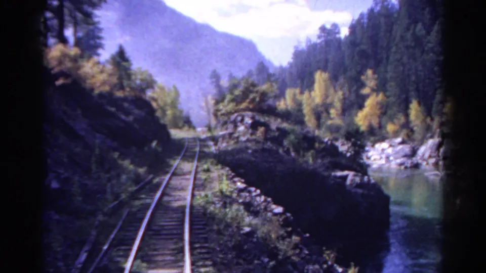 1967 Colorado mountain rail view with tracks curving along a river, dark rock banks, evergreens, yellow foliage, and distant hills.