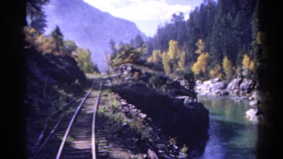 Rear-facing 1967 Colorado train frame with tracks, blue-green river water, rocks, evergreens, autumn trees, and hazy mountains.