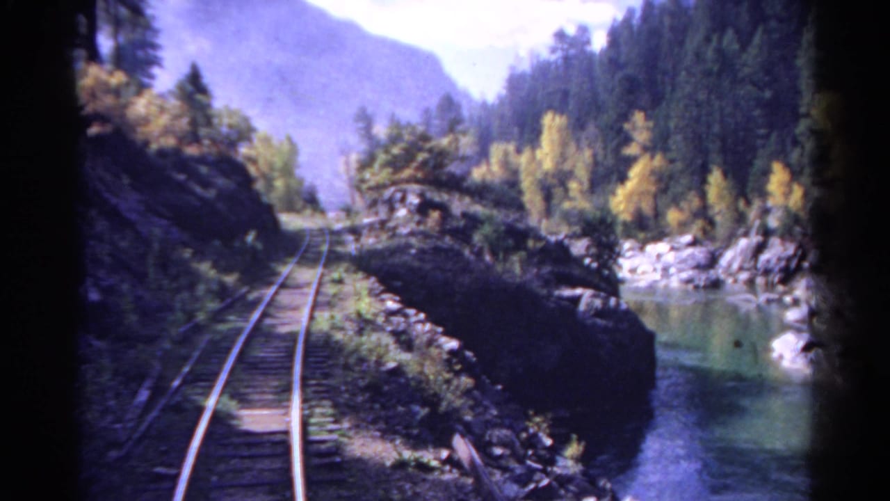 1967 Colorado 8mm frame showing a curving railroad track along a rocky river with forested slopes and distant mountains.