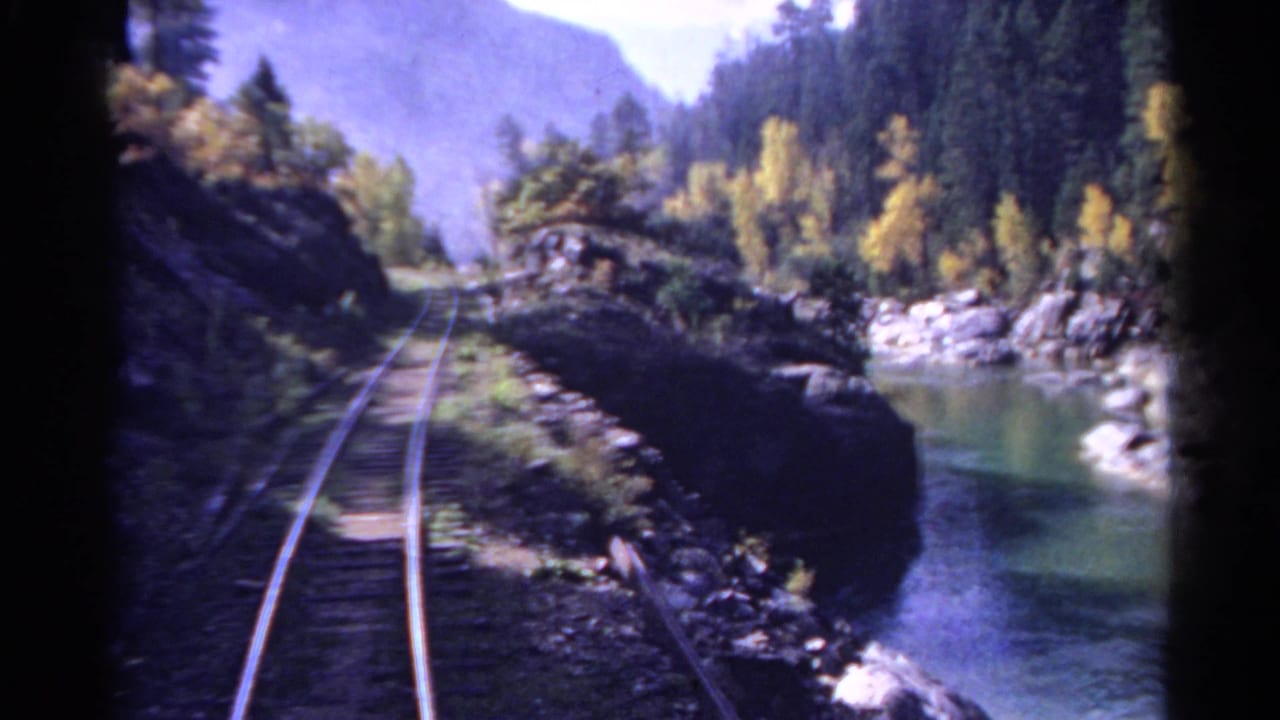 1967 Colorado train-view frame with rails bending along a narrow river corridor lined by rocks, evergreens, and yellow trees.