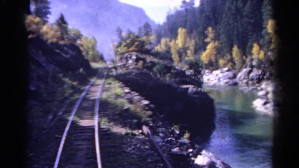 1967 Colorado train-view frame with rails bending along a narrow river corridor lined by rocks, evergreens, and yellow trees.