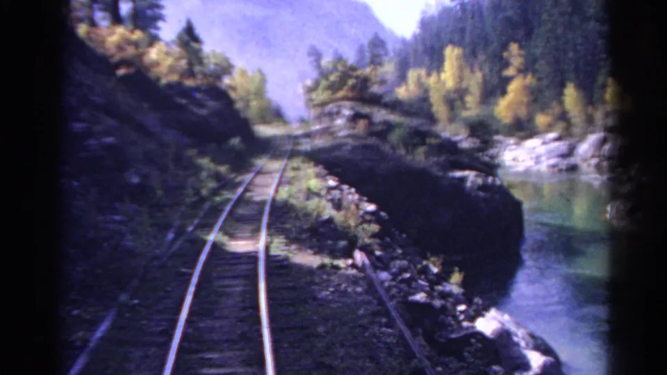 1967 Colorado 8mm frame looking down a riverside rail line with rocks, green water, trees, and hazy mountain forms beyond.
