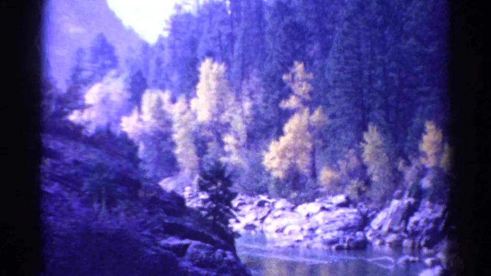 1967 Colorado 8mm frame showing a rocky river, dense evergreen trees, yellow foliage, and purple-toned mountain light from the train route.