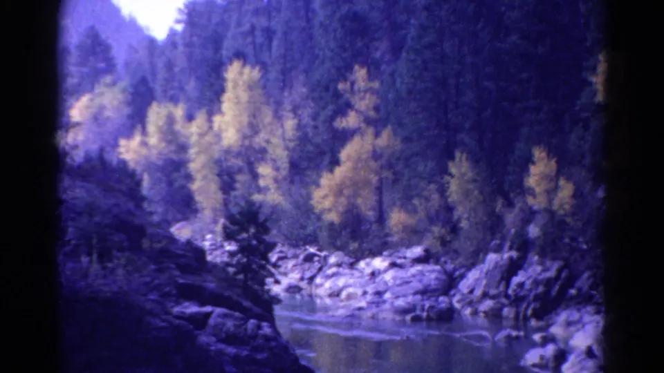 Soft 1967 Colorado 8mm view of a river pool bordered by rocks, yellowing trees, dark conifers, and shadowed valley slopes.