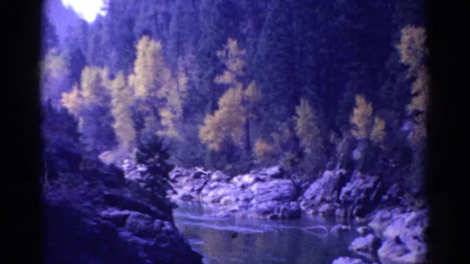 1967 Colorado 8mm frame with calm water, pale rocks, yellow foliage, and dense evergreen forest inside dark rounded film borders.