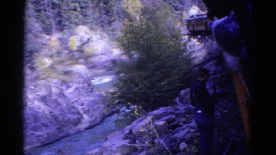1967 Colorado 8mm frame showing a person on a rocky ledge above a narrow river and wooded canyon.
