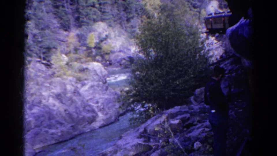 1967 Colorado home-movie frame with a person at the right edge looking across a rocky river gorge and forested slope.