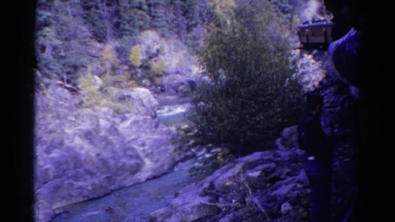 1967 Colorado 8mm frame showing a traveler by rocks above a river, with trees, canyon stone, and part of a train at the side.