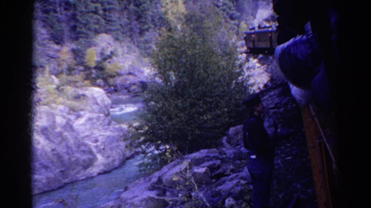 1967 Colorado home-movie frame of canyon rocks, river water, trees, and a partially visible train beside a dark-clad traveler.