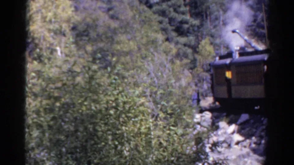 1967 Colorado home-movie frame of a steam train passing through a forested rocky canyon, partly obscured by trees.
