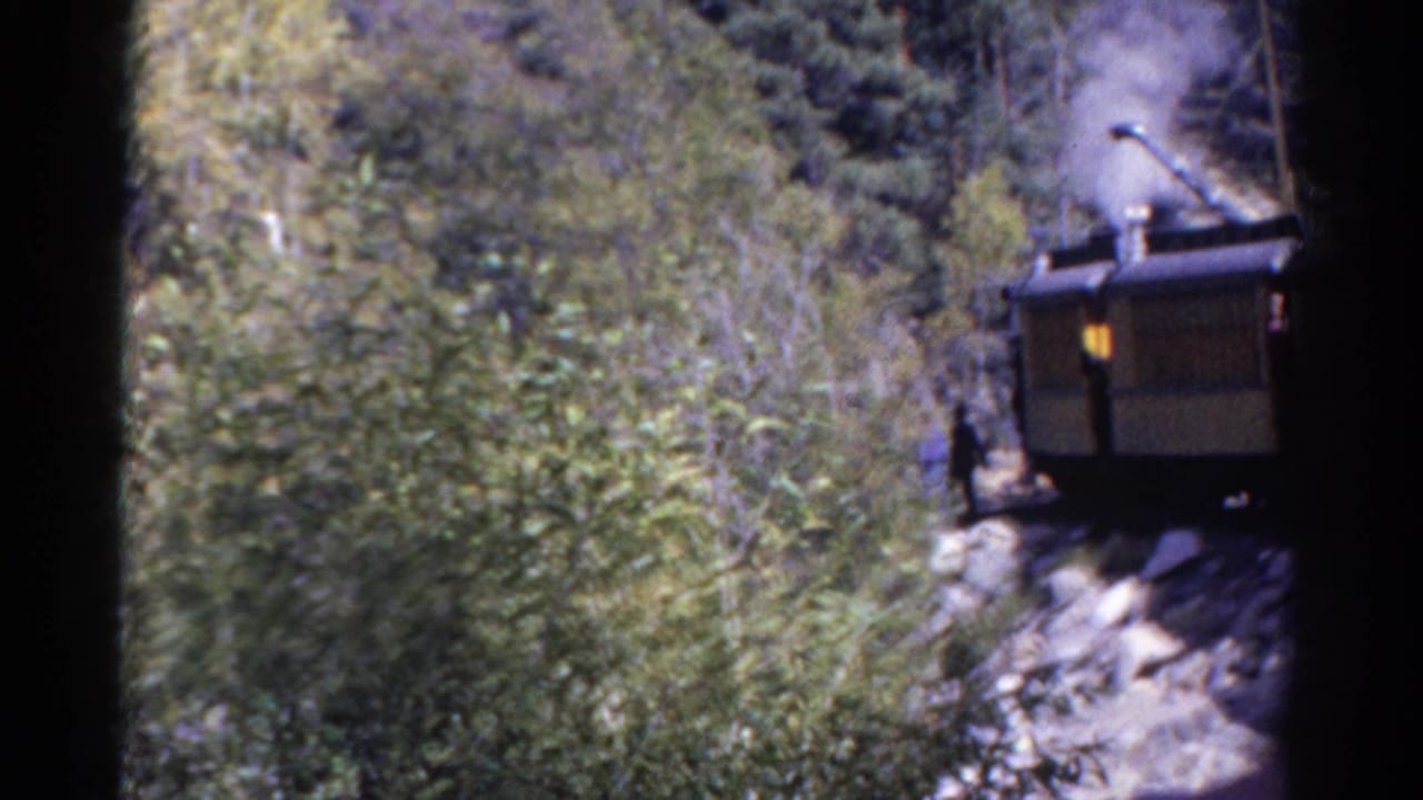 Steam rail machinery partly seen beside trees and rocks, with smoke rising and dark frame edges.