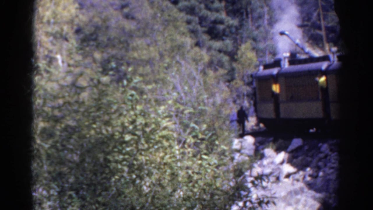 1967 Colorado home-movie frame with a man beside steam-powered rail machinery in a wooded rocky pass.