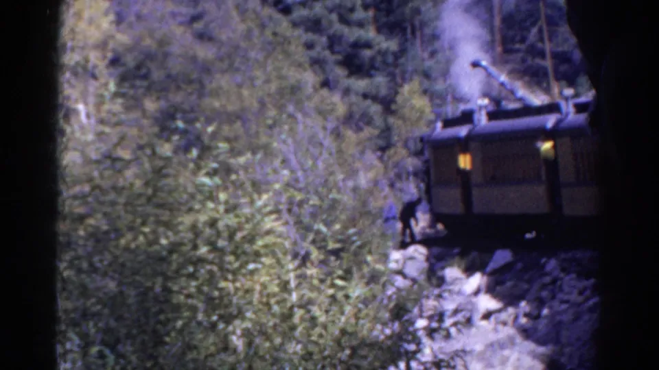1967 Colorado frame showing a man beside steam-powered rail machinery near boulders and dense mountain trees.