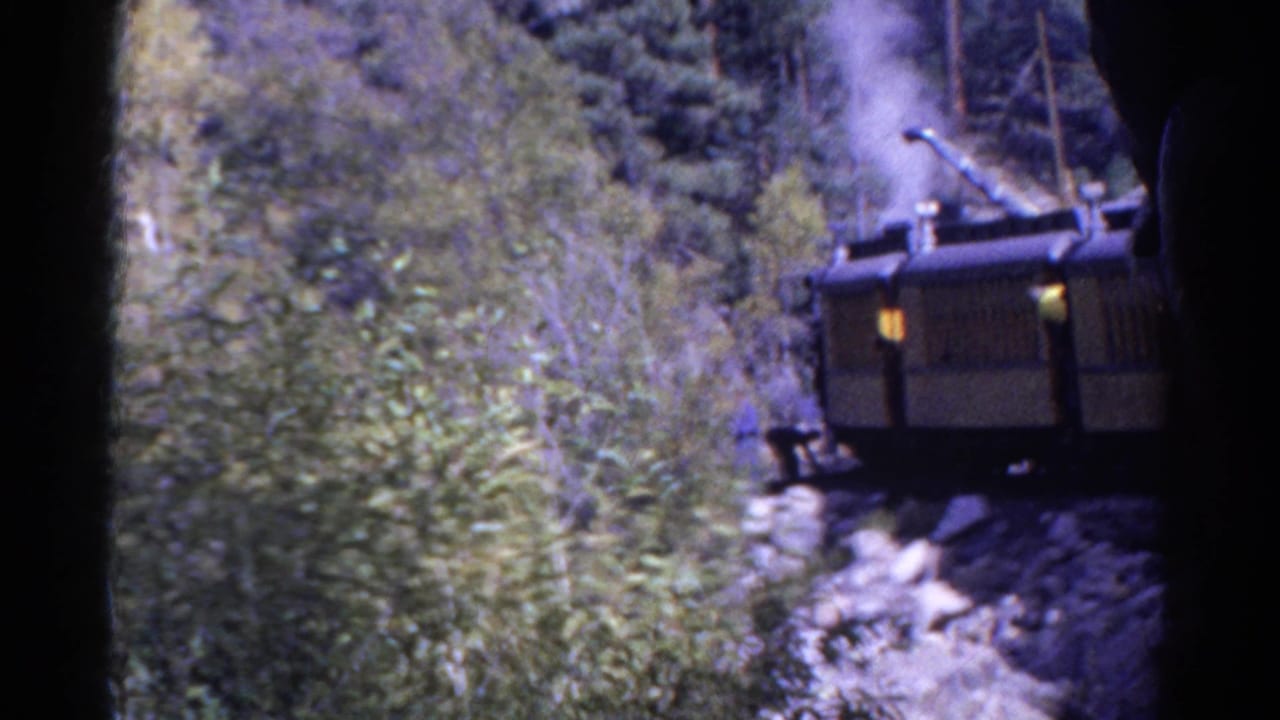 1967 Colorado 8mm frame showing a steam train partly obscured by brush and trees on rocky mountain terrain.