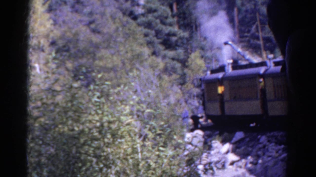 1967 Colorado home-movie frame with a steam train emitting smoke, partly hidden by forest growth on a mountain slope.