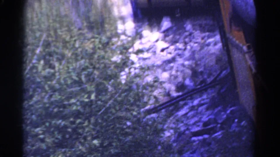 1967 Colorado home-movie frame of a rocky, brush-lined riverbank, with the train no longer clearly visible.