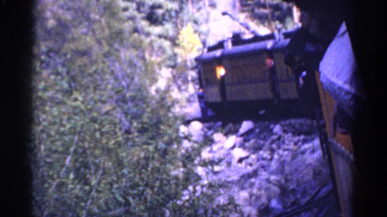 Derailed rail car beside rocky trackside foliage, seen from a passing train.