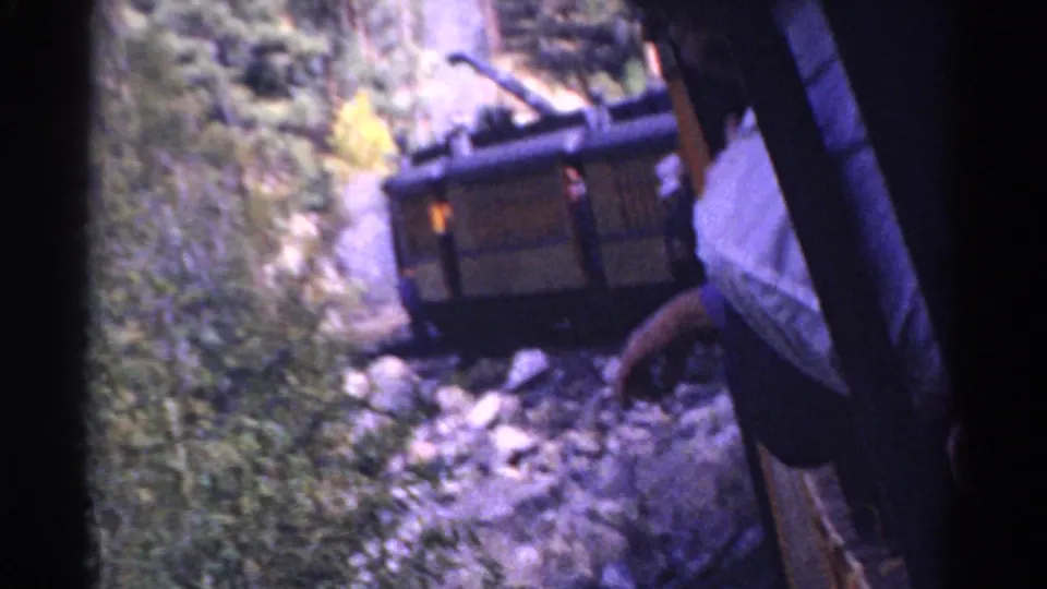 1967 Colorado 8mm frame with a passenger in a light shirt at the train edge, looking toward a derailed rail car in rocky foliage.