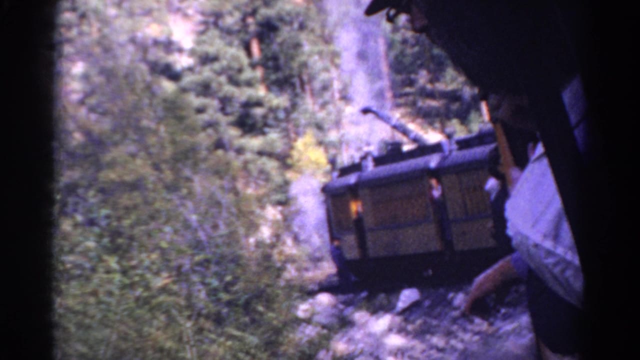 1967 Colorado 8mm frame showing a wooden rear cart on a steep mountain railway, with trees, rocks, and a nearby passenger partly visible.