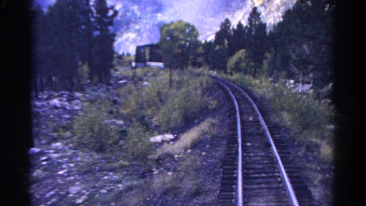 1967 Colorado 8mm frame looking from a moving train toward curving tracks, pine trees, rocks, and a small bridge ahead.