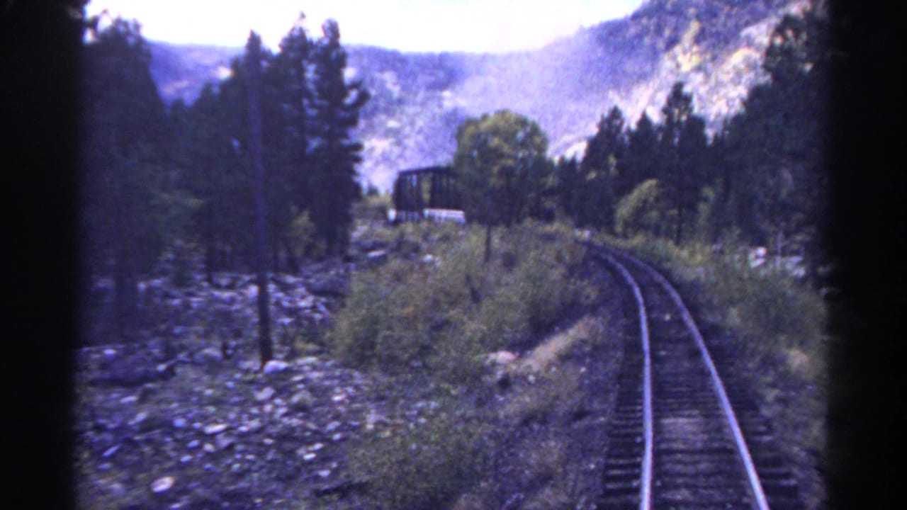 1967 Colorado 8mm frame of a mountain rail line curving past pine trees and scrub toward a bridge-like structure.