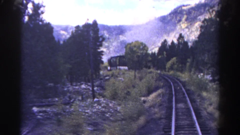 1967 Colorado 8mm frame showing a train-window view along curved track through wooded rocky mountain scenery.