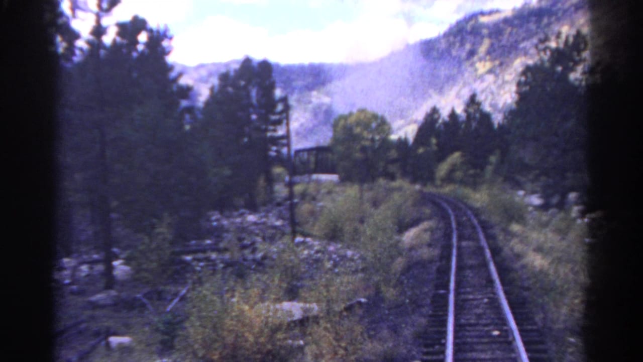 1967 Colorado train-view frame showing curving tracks, pine forest, rocky ground, and mountains ahead.