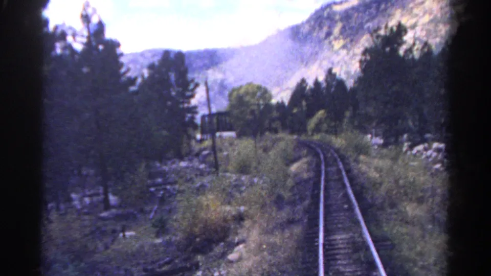 1967 Colorado train-view frame with rails bending through pine forest, brush, rocks, and mountain slopes.