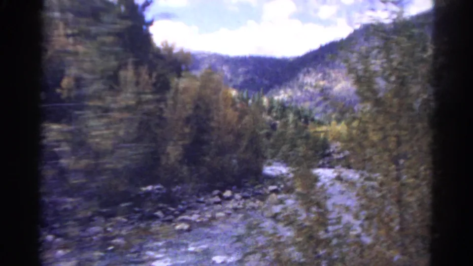1967 Colorado train-view frame with a rocky riverbank, autumn foliage, pine trees, and distant mountains.