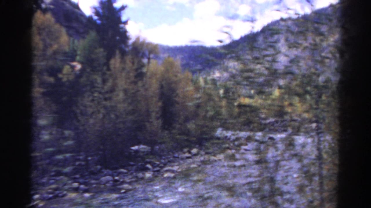 1967 Colorado 8mm frame showing a shallow rocky river, evergreens, yellow foliage, and blurred mountain slopes from a moving vehicle.