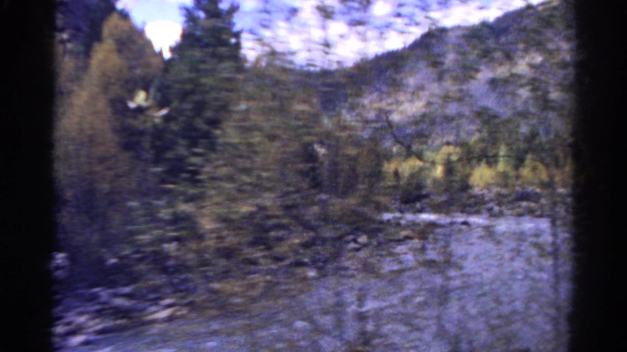 1967 Colorado home-movie frame with motion-blurred trees, a shallow river, rocky banks, and forested mountain terrain.