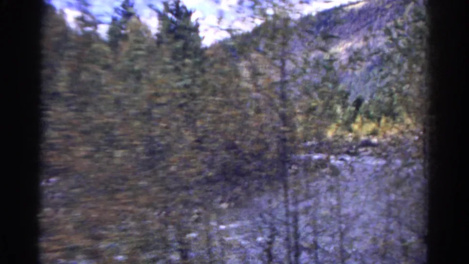 1967 Colorado home-movie frame showing a shallow river, dense blurred trees, autumn foliage, and rocky mountain slopes.