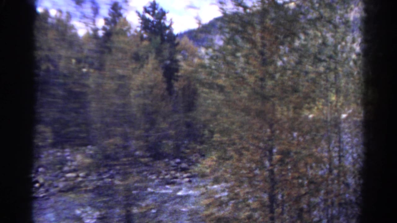 Blurred train-window view of a rocky mountain river and trees with dark film edges.