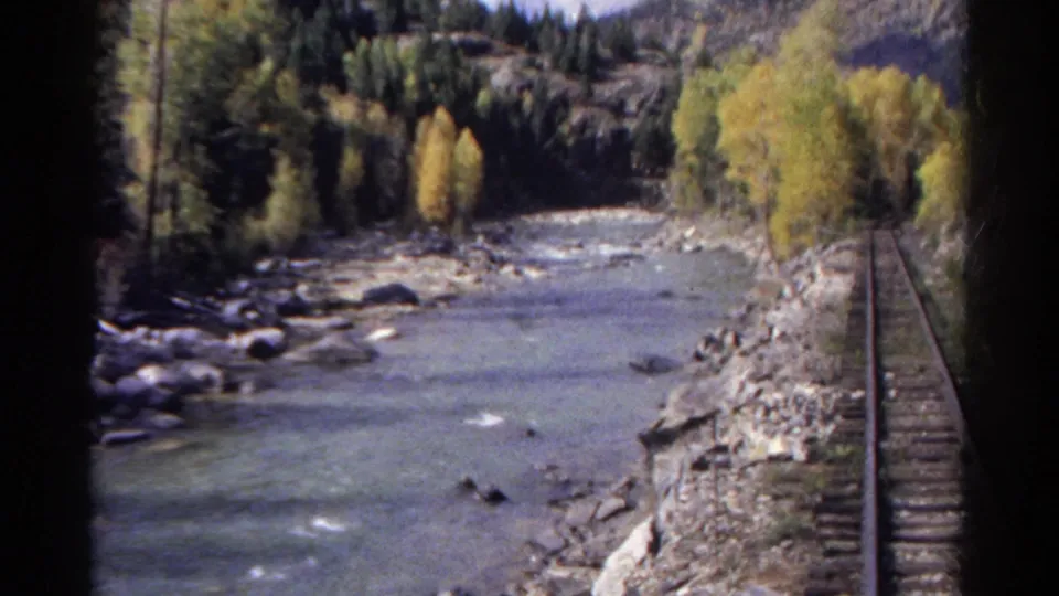 1967 Colorado 8mm view from a moving train, with rails beside a shallow river, rocky banks, and autumn trees.