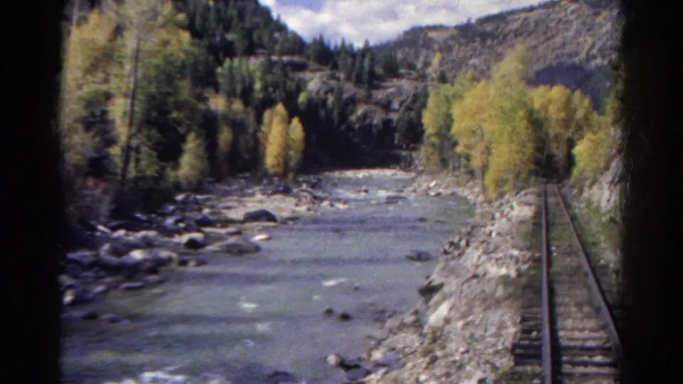 1967 Colorado home-movie frame showing railway tracks beside a shallow river, autumn trees, rocks, and forested slopes.