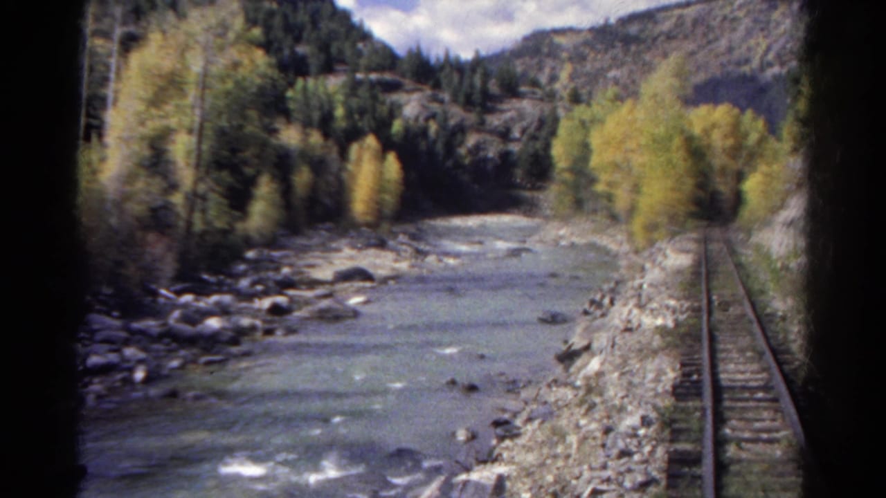 1967 Colorado 8mm view from a moving train beside a rocky river, railroad track, trees, and mountain slopes.