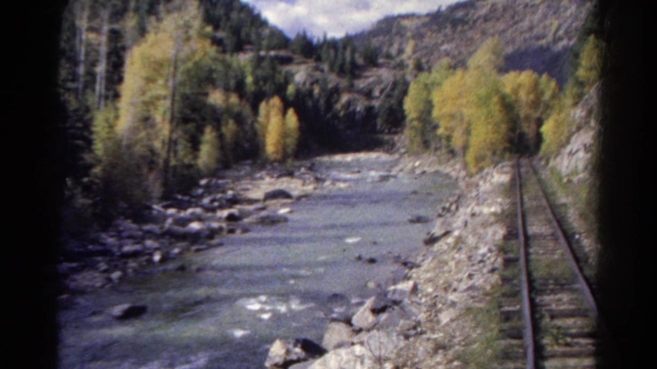 Vintage Colorado train-view frame showing a single track, river rocks, autumn-colored trees, and forested ridges.