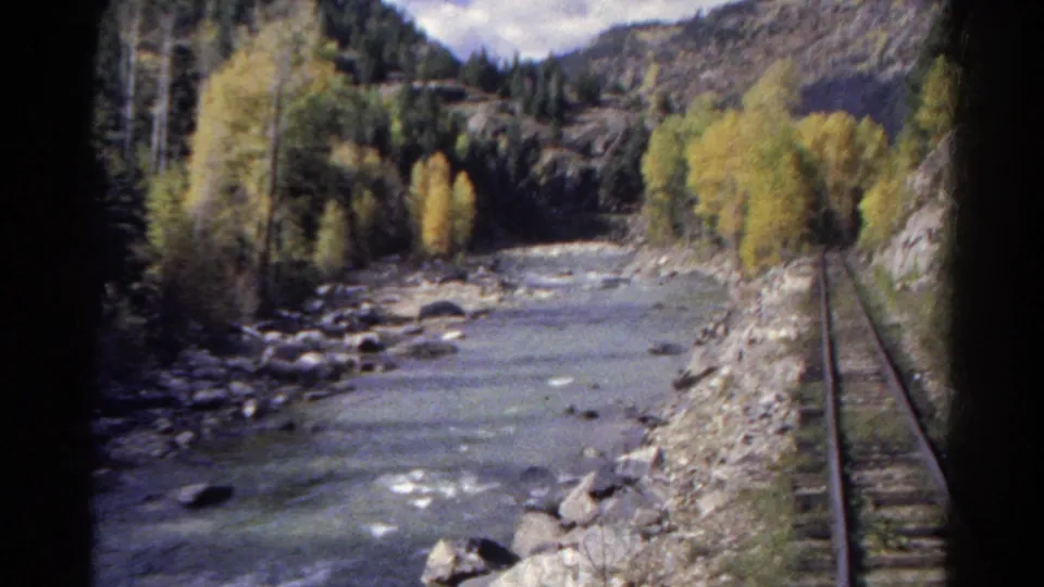 Vintage Colorado train-view frame showing a single track, river rocks, autumn-colored trees, and forested ridges.