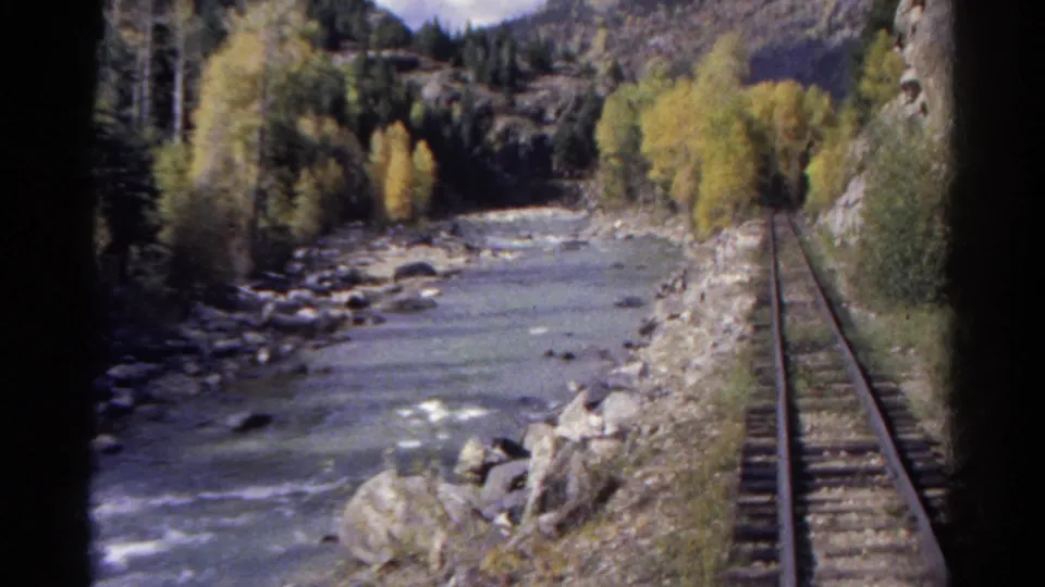 1967 Colorado home-movie frame with a rocky river bend, parallel railroad track, trees, and steep valley edge.