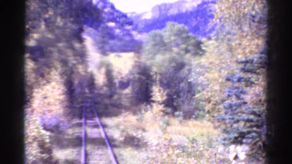1967 Colorado home-movie frame showing a narrow railway track running through trees toward a bright mountain valley.