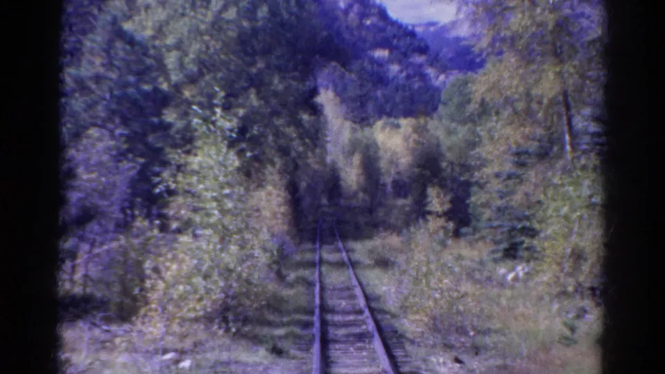 1967 Colorado train-window view of rails, close forest growth, distant ridges, and cloudy sky in grainy 8mm color.