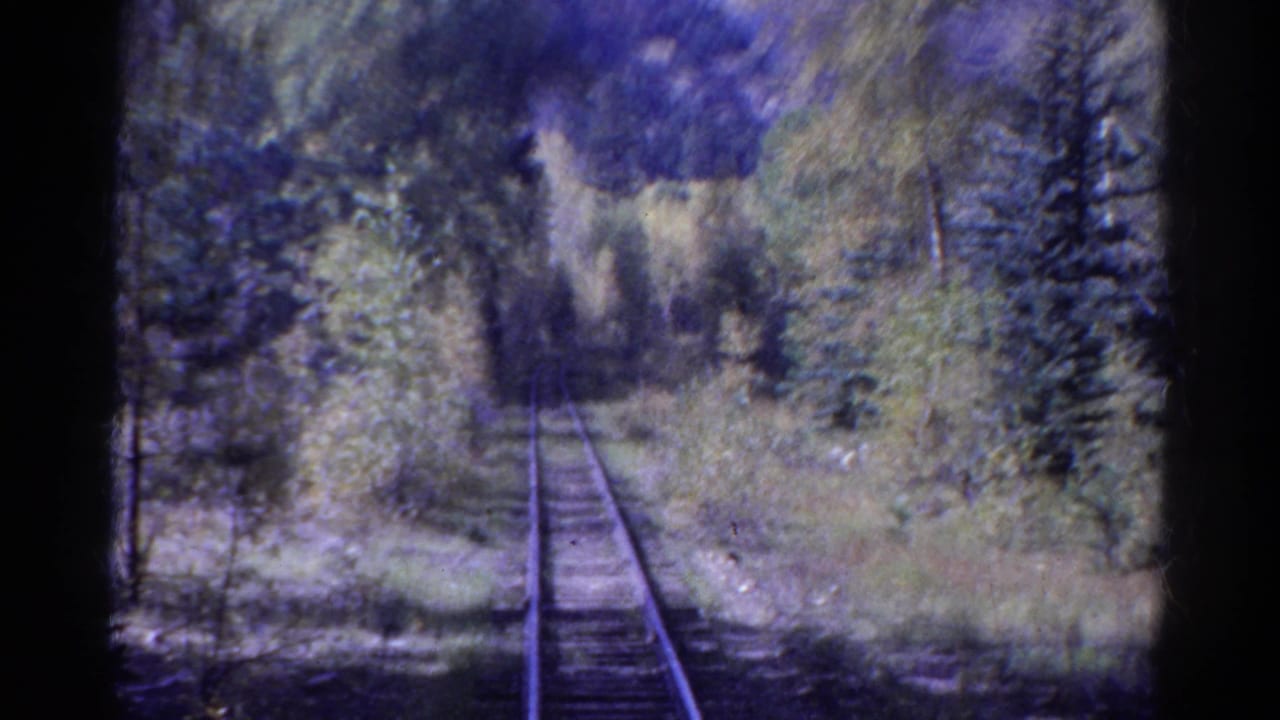 1967 Colorado rail-view frame with tracks, wooden ties, dense trees, pale brush, and soft home-movie blur.