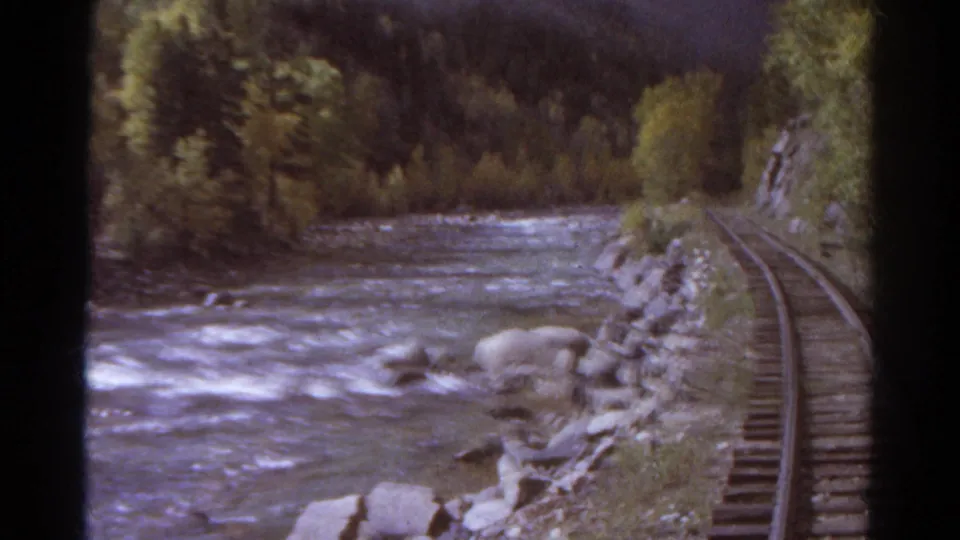 1967 Colorado 8mm frame showing a rushing river beside a railway line, with rocks, trees, and wooded slopes along the bank.