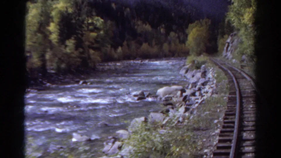 1967 Colorado 8mm frame of railway track beside white water, with rocky banks, trees, and shaded mountain terrain.