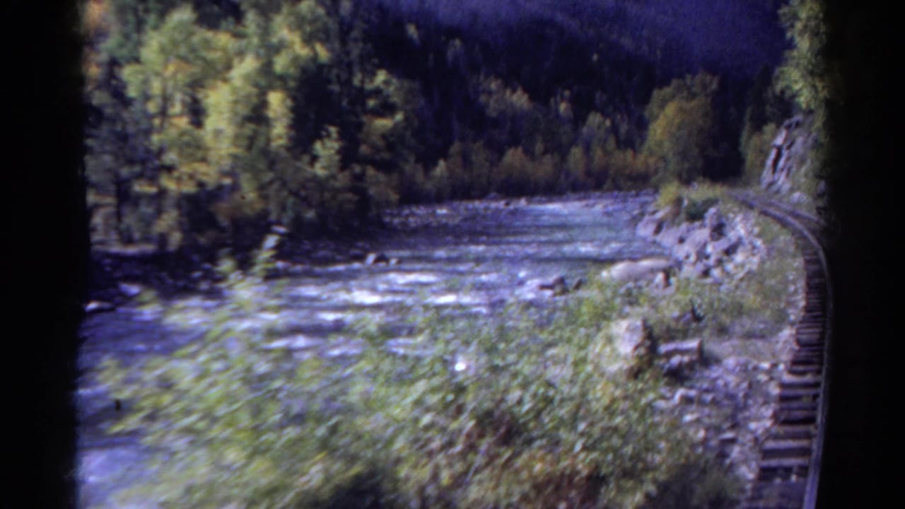 1967 Colorado 8mm frame with motion-blurred brush in the foreground, river rapids, rocks, and curving track at right.